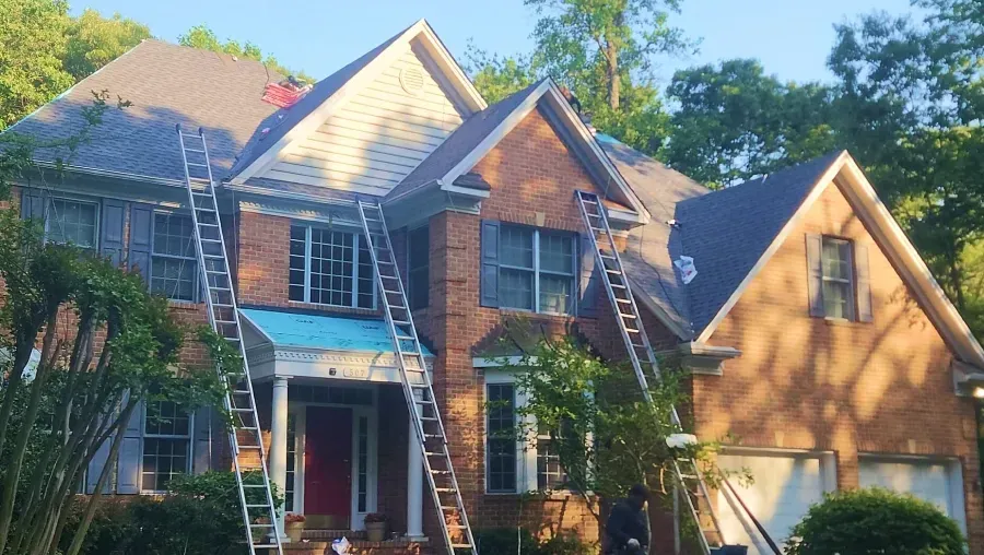 Two-story brick and wood house with ladders on the front, surrounded by trees under a bright blue sky