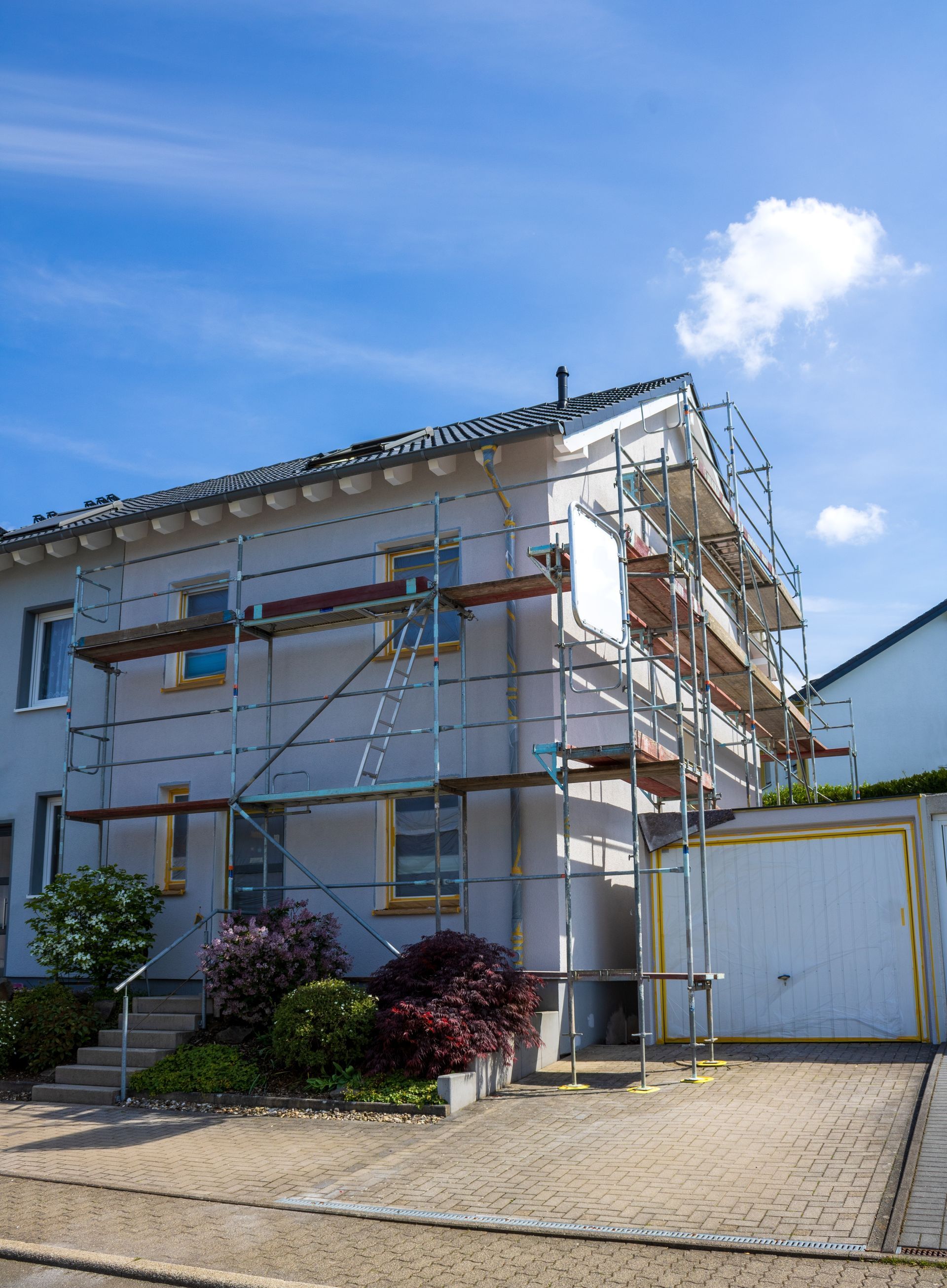 Two-story house under renovation with scaffolding, white exterior, and a driveway under a blue sky