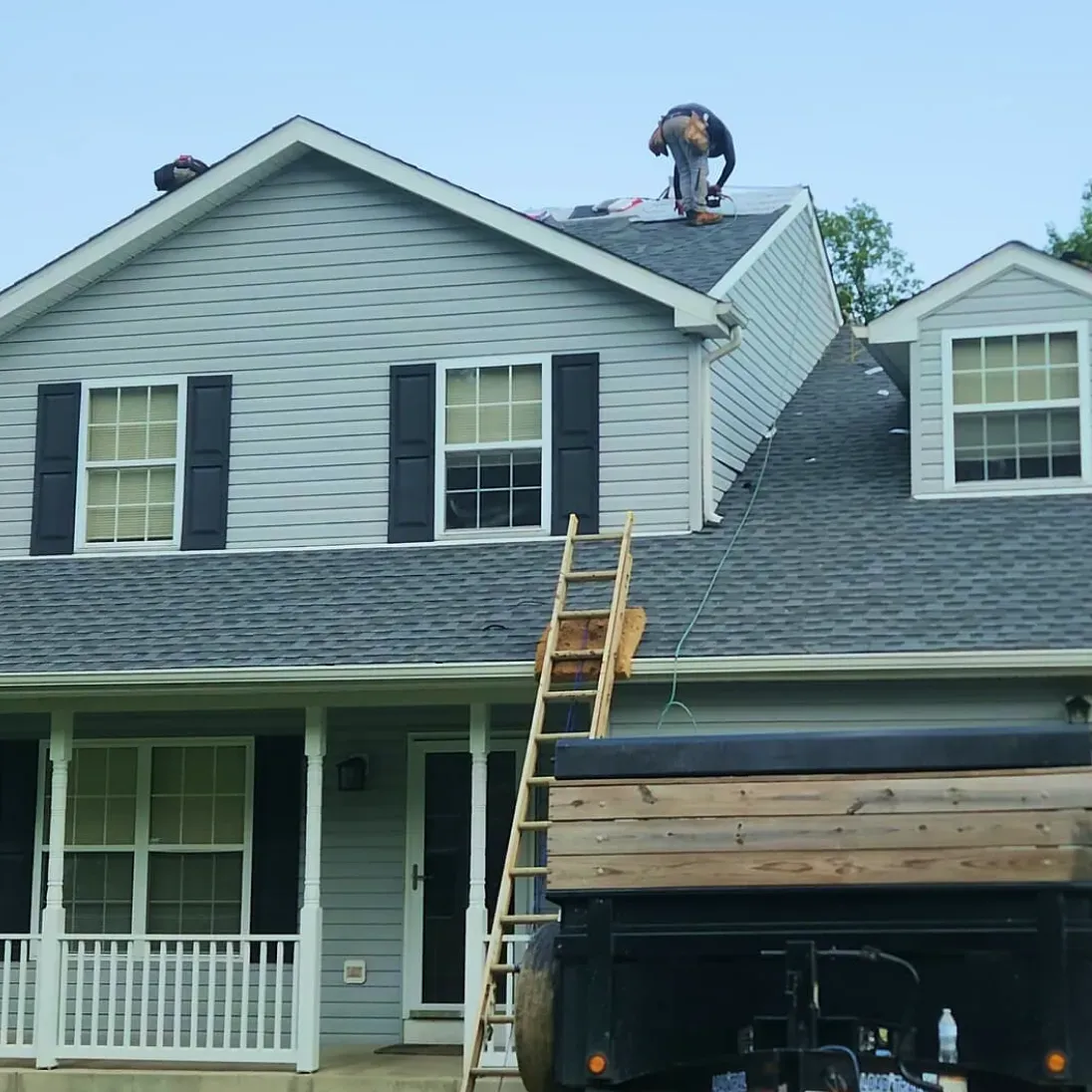 Worker on a roof beside a ladder, with a house front and garage visible below.