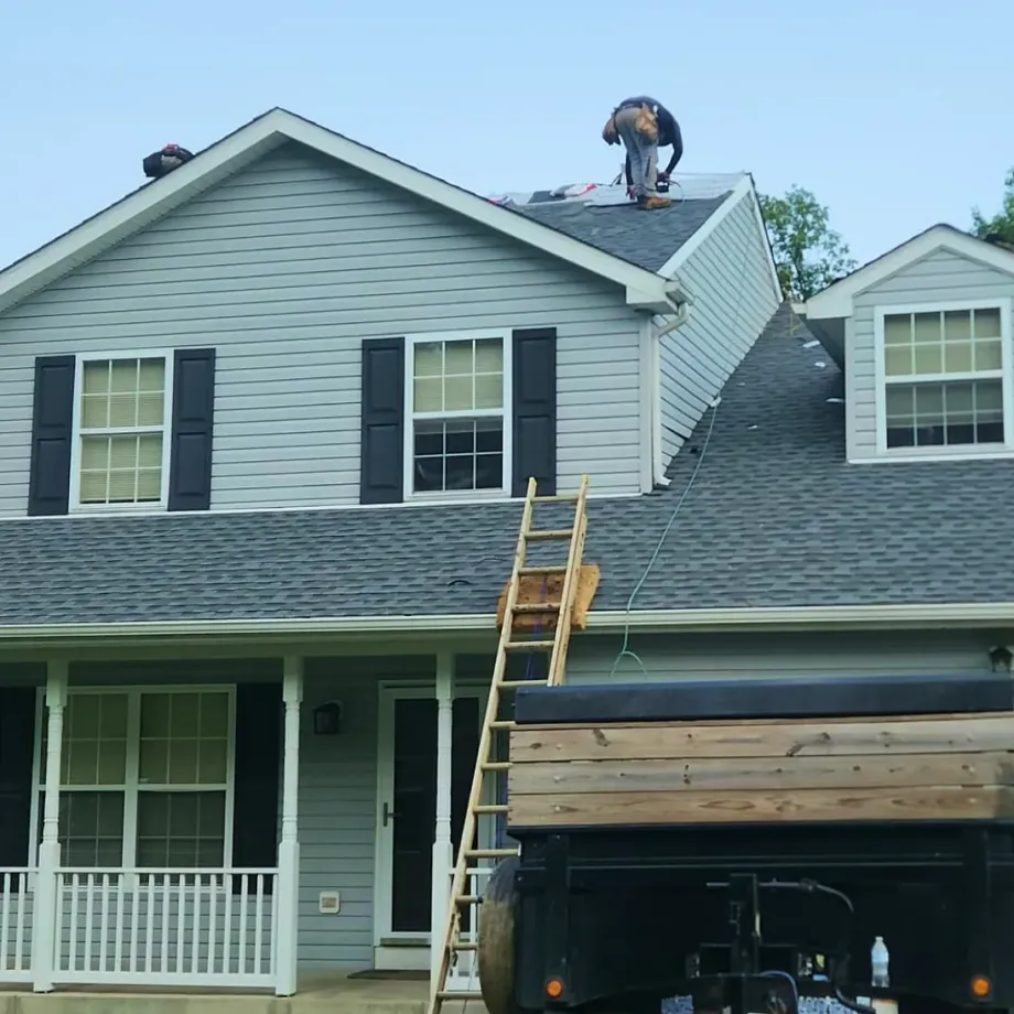 Worker on a roof beside a ladder, with a house front and garage visible below.