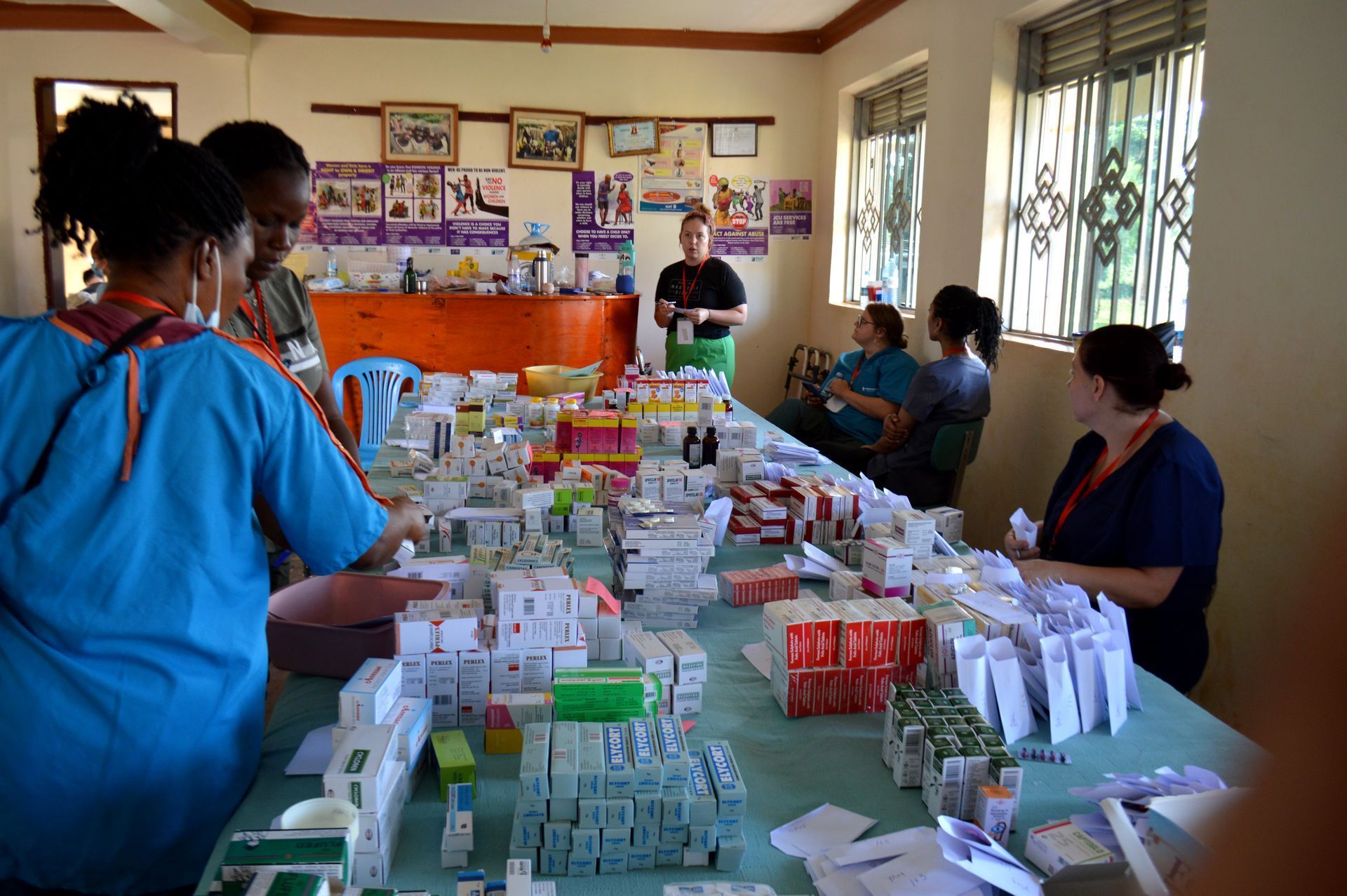 People sorting medication packages on a table in a clinic. Brightly lit room with a window.