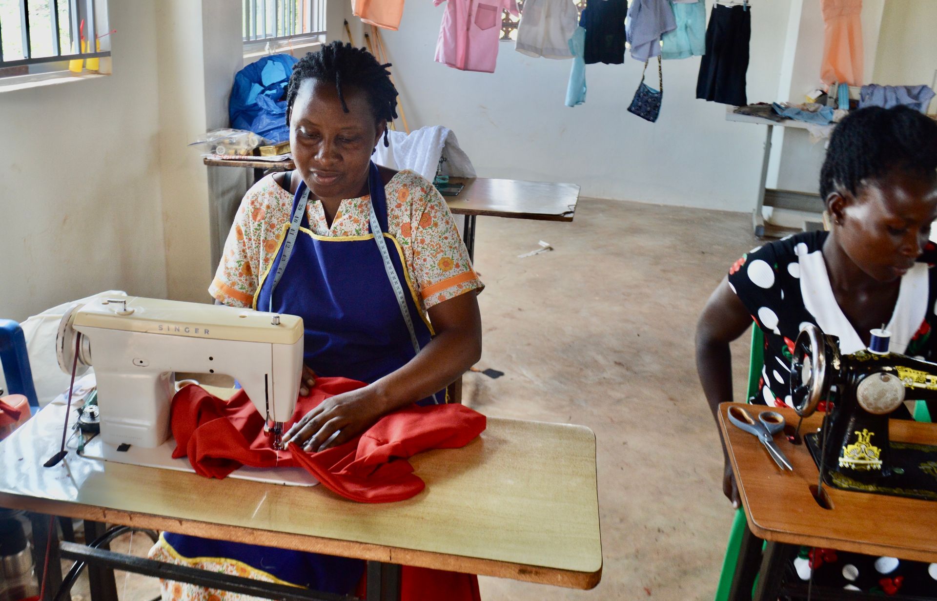 Two women sewing red fabric in a workshop. Clothesline with clothes in the background.