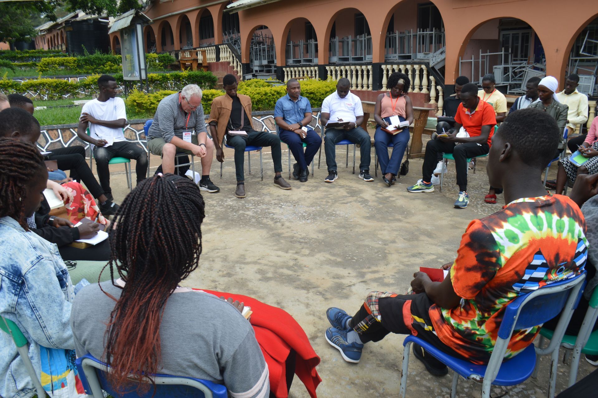 Group of people in a circle, discussing. Outdoor setting. Some taking notes.