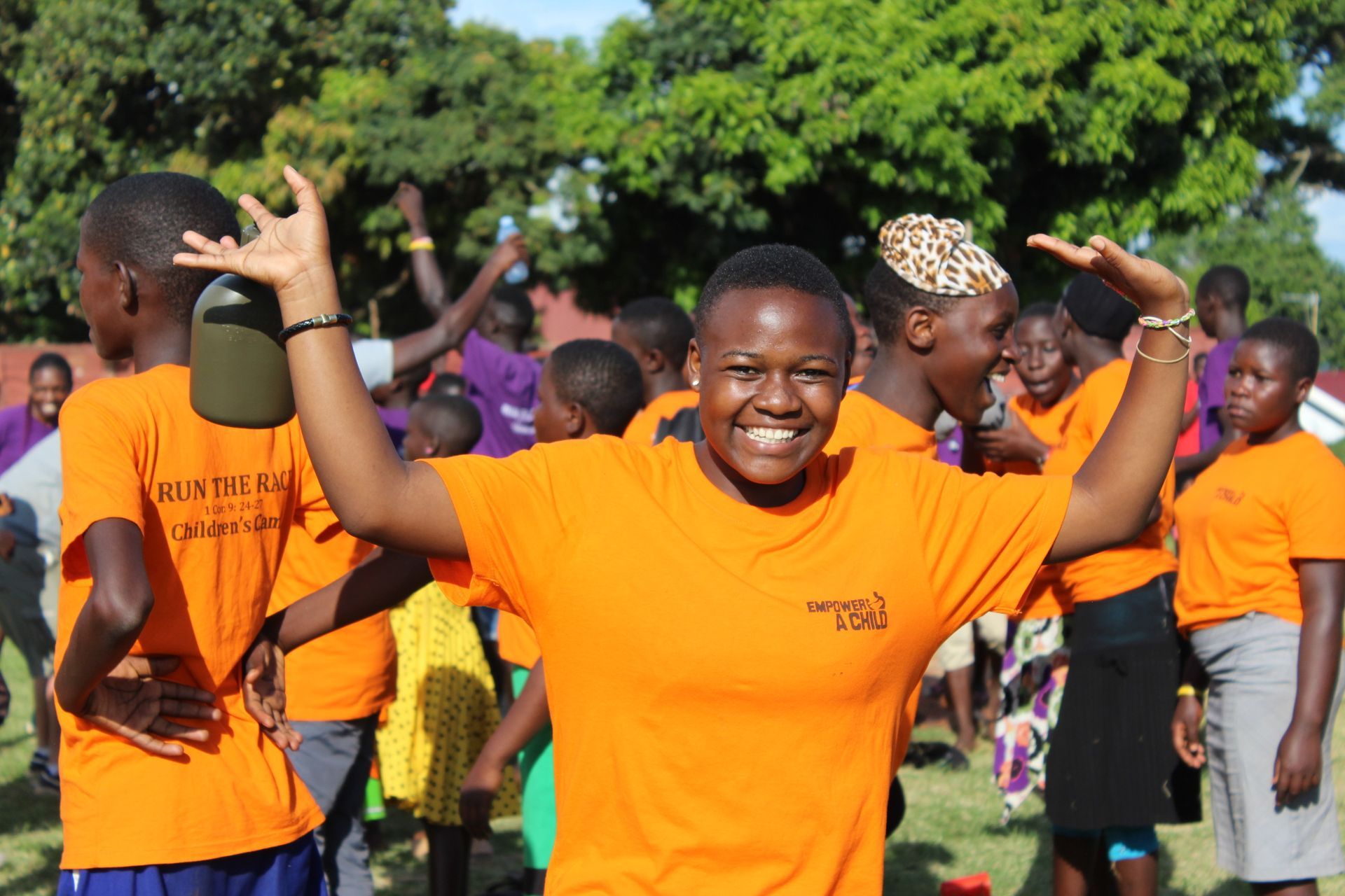 Woman in orange shirt with arms raised smiling in a group of children, outside.