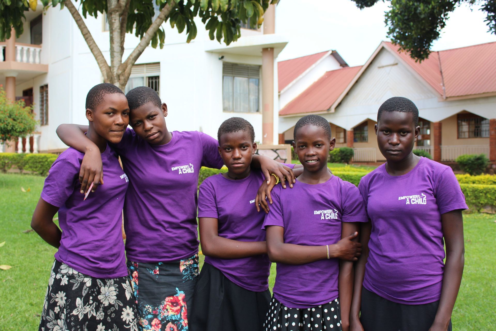 Five girls in purple shirts pose outside a building; they are smiling, some with arms around each other.