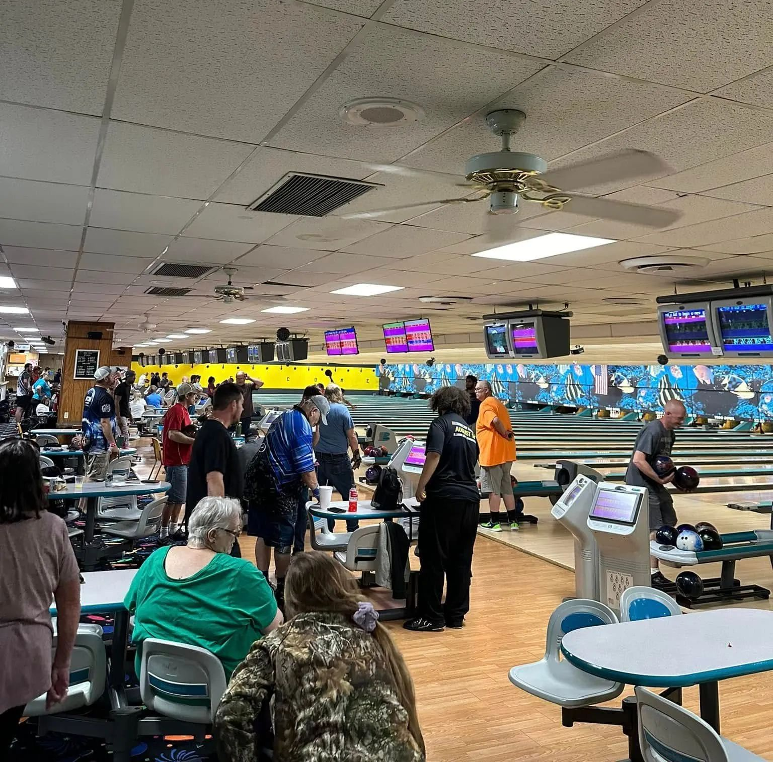 Crowded bowling alley with players on the lanes and spectators seated nearby under bright scoreboards