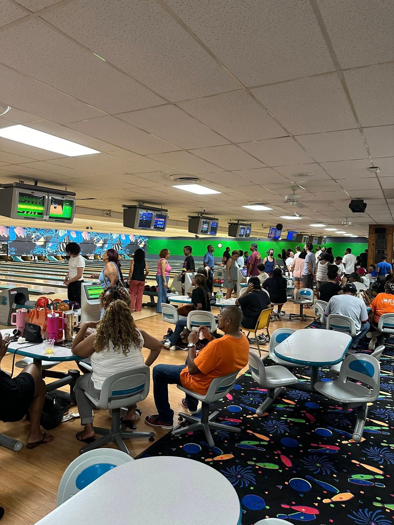 Busy bowling alley with people seated at lanes and glowing score screens overhead