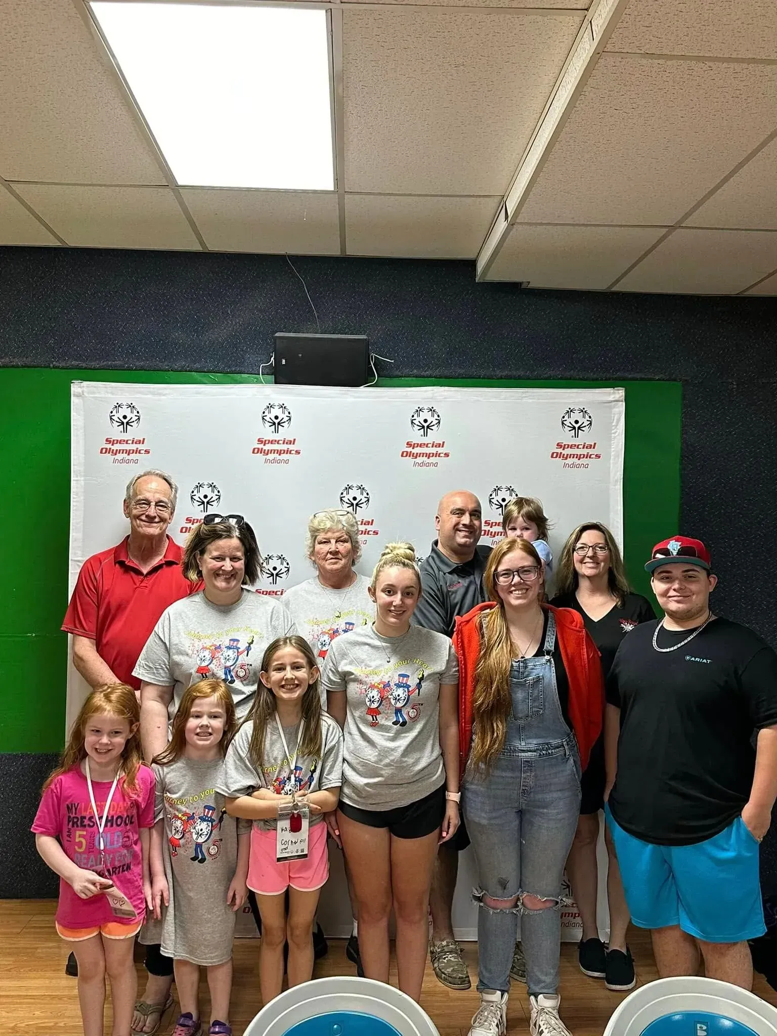 Group of diverse people smiling in front of a backdrop with logos. Some are children. Indoors.