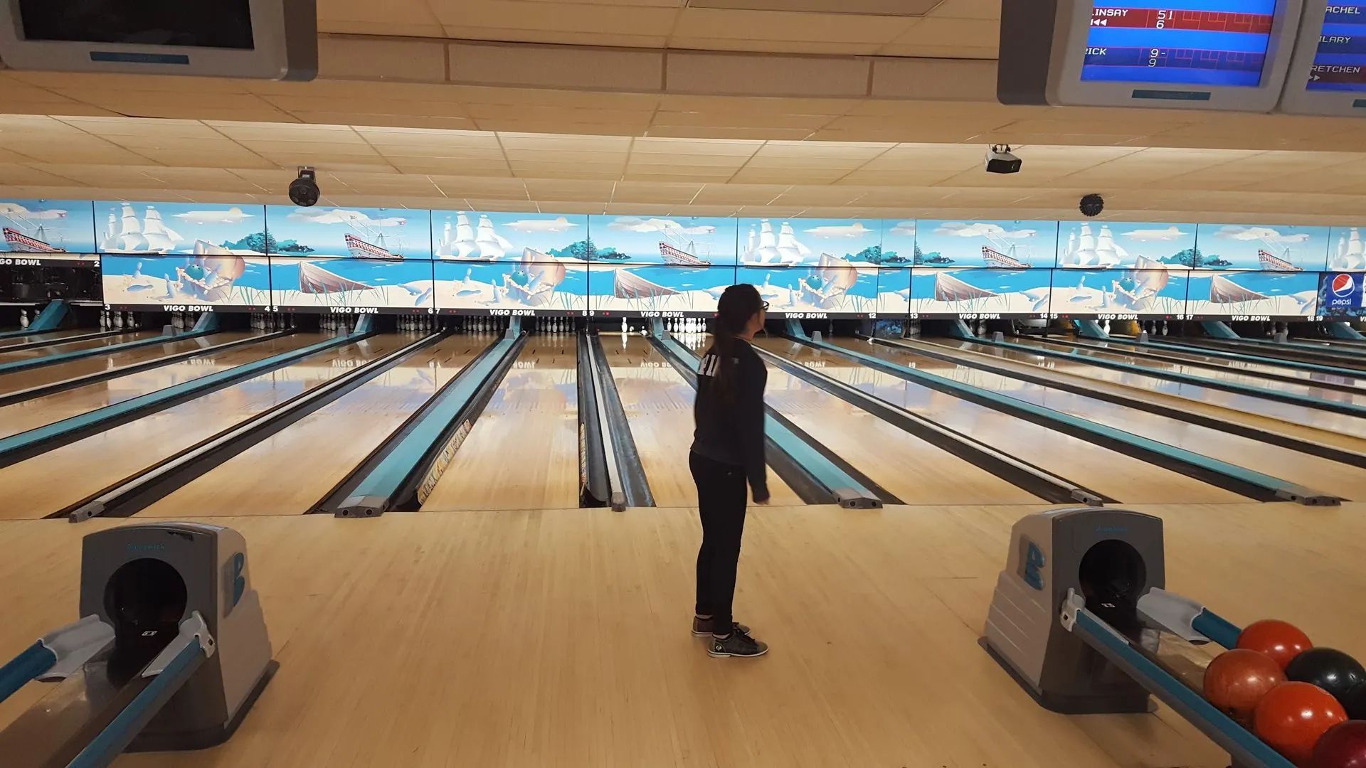 Bowling alley with a person standing at the lane, bowling pins lit on the far end, and balls in the rack.