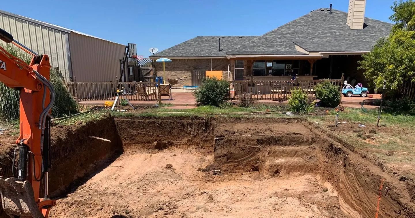 Excavated pool hole in a backyard, with an excavator on the left and a house in the background.