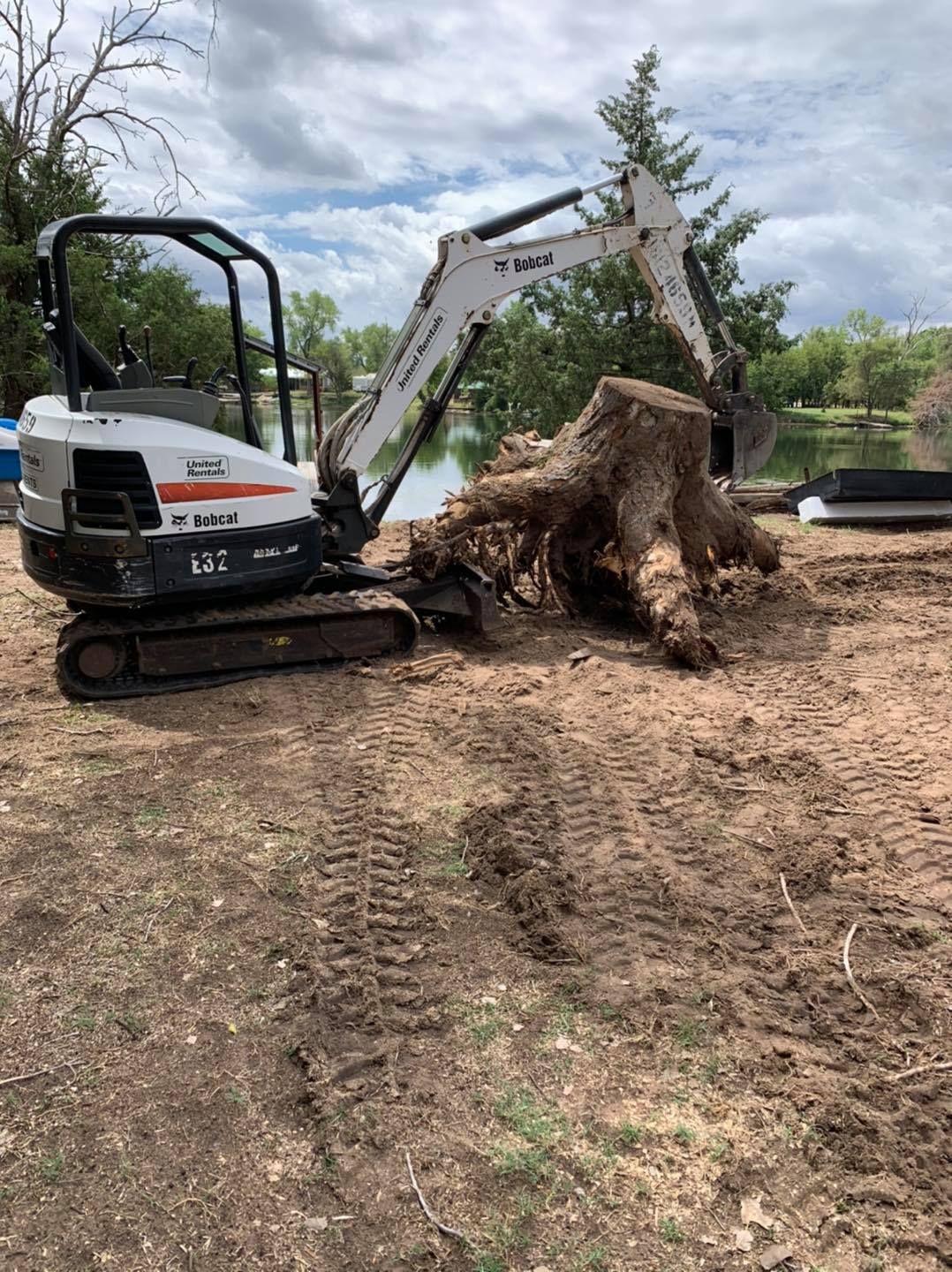 Bobcat excavator removing a tree stump on dirt near water, under a cloudy sky.