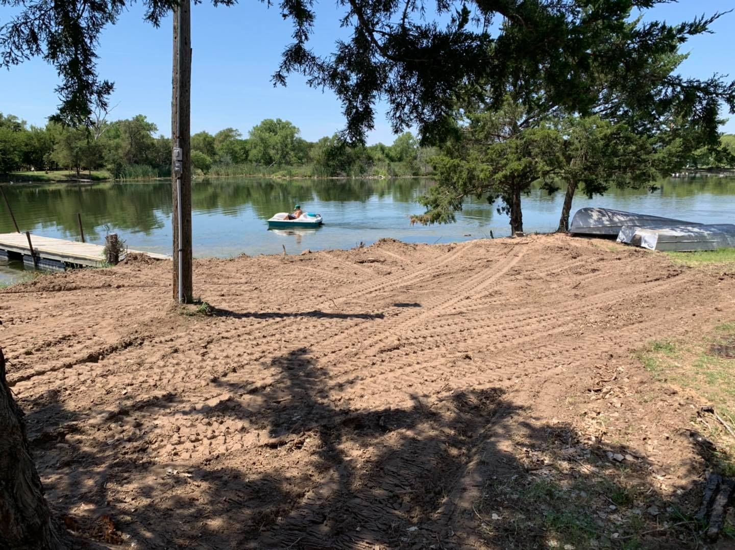 Lakeside view with a newly tilled area. A small boat floats on the water. Trees provide shade and frame the scene.