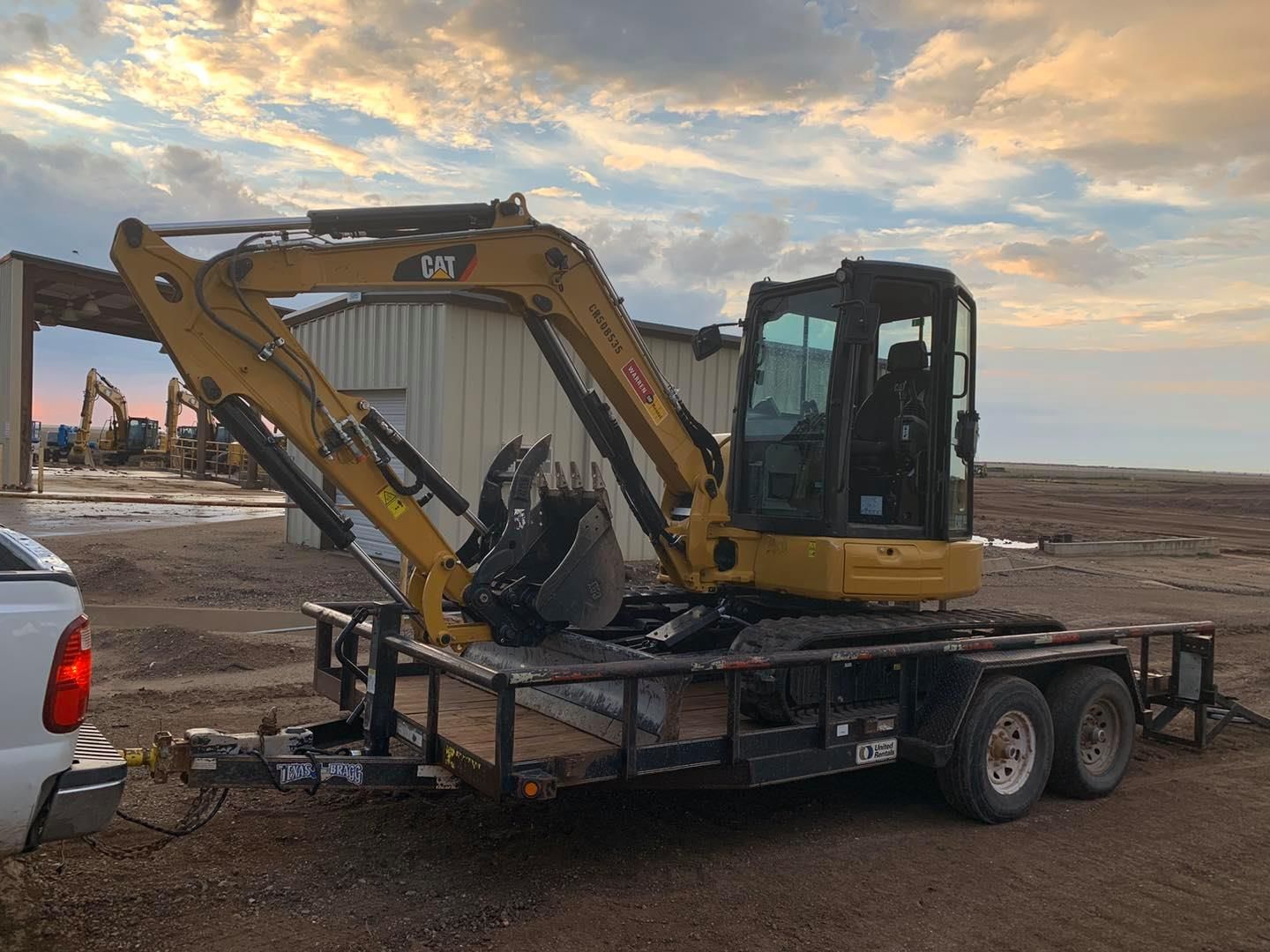 Yellow CAT excavator on a trailer hitched to a white truck, in a construction site at dusk.