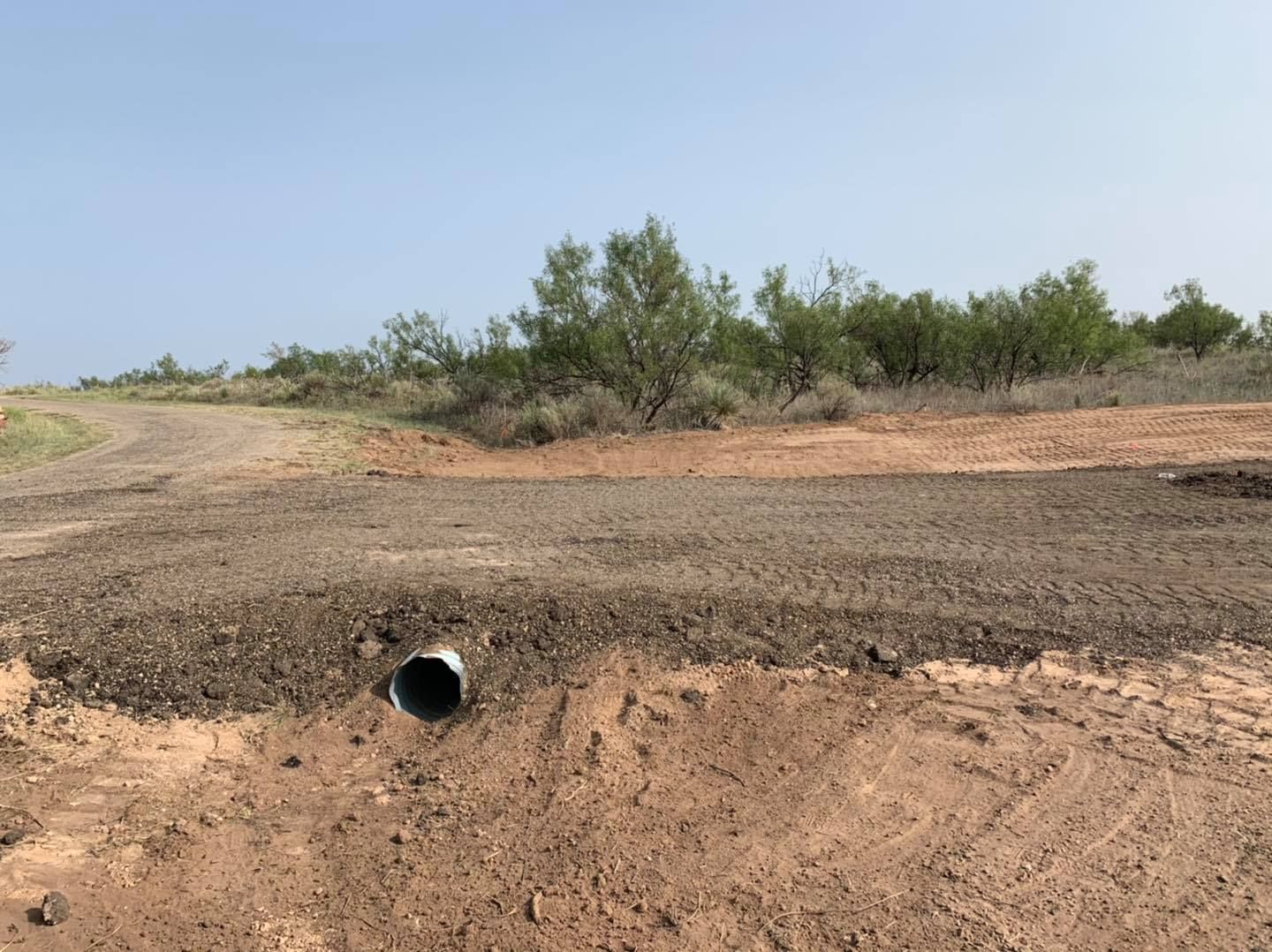 A culvert pipe drains a dirt road, with sparse vegetation under a blue sky.