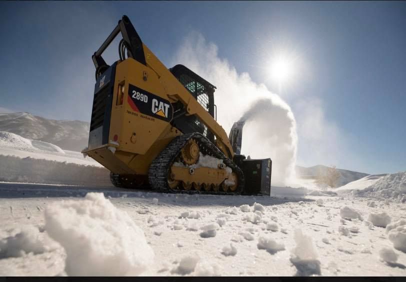 Yellow Caterpillar skid steer snow blowing on a snow-covered road under a bright sun.