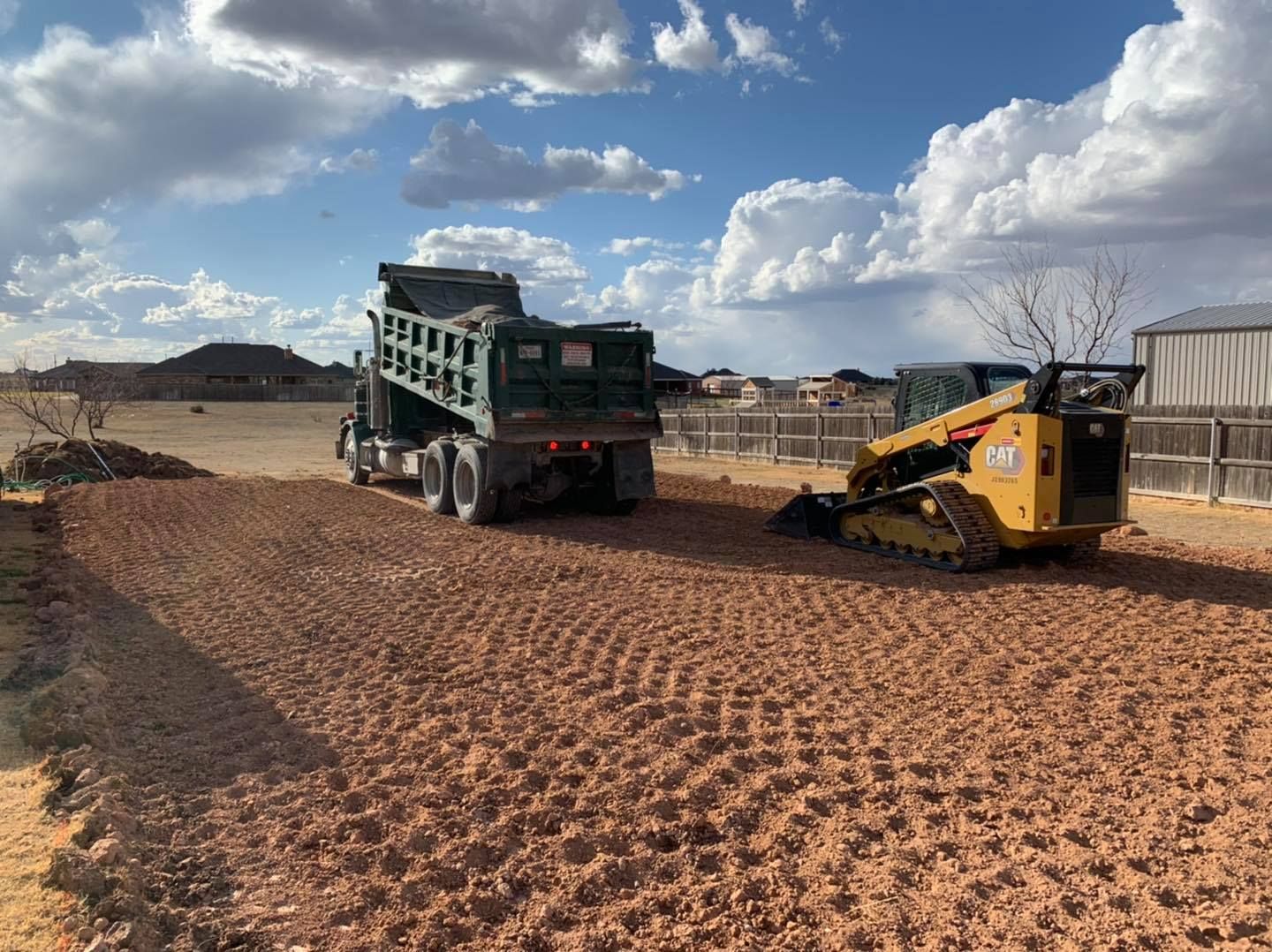 A dump truck unloading material next to a yellow skid steer on a dirt area under a blue sky.