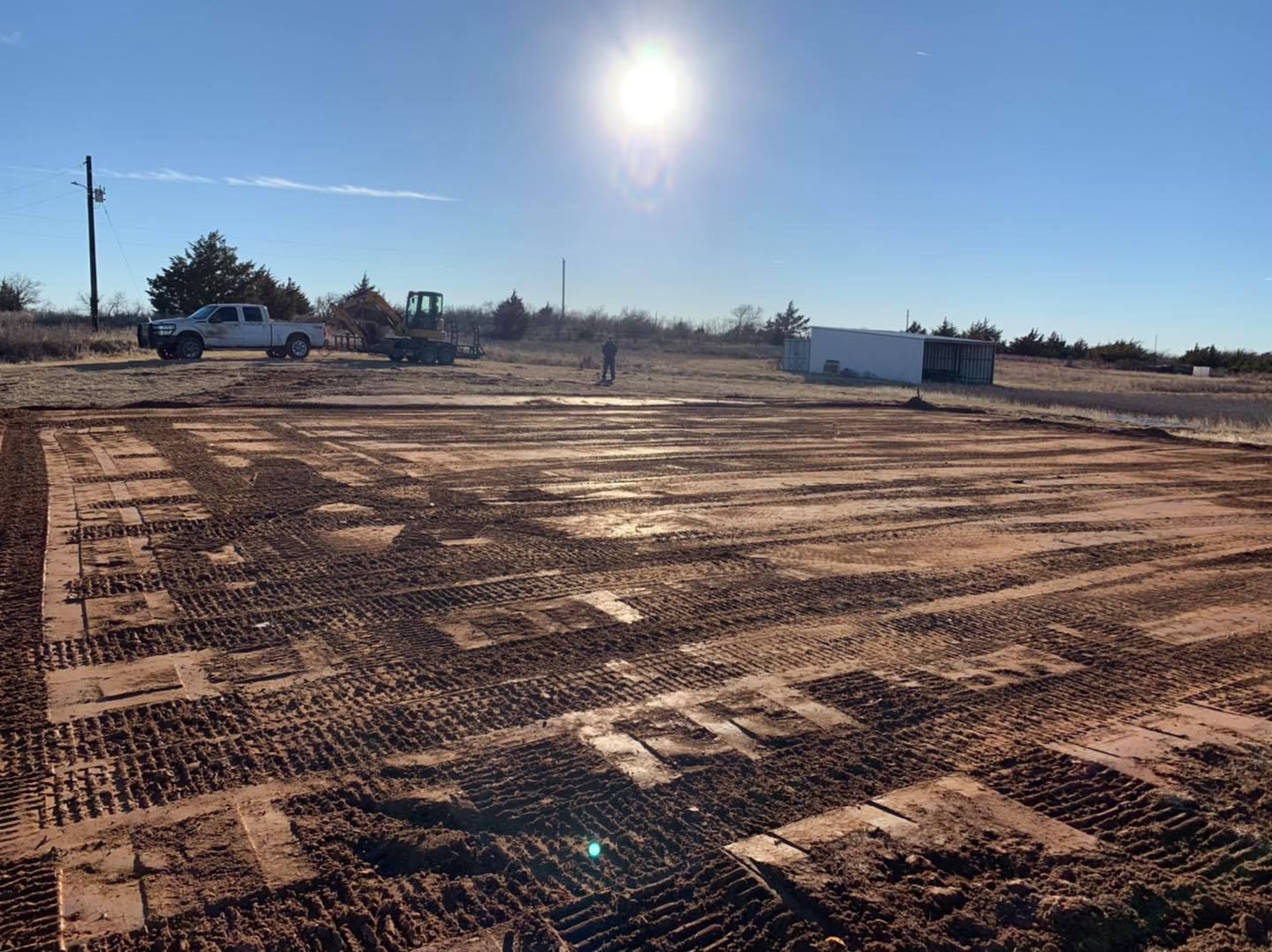 Construction site, sunlit with graded dirt, possibly for a foundation. Vehicles and equipment in the background.