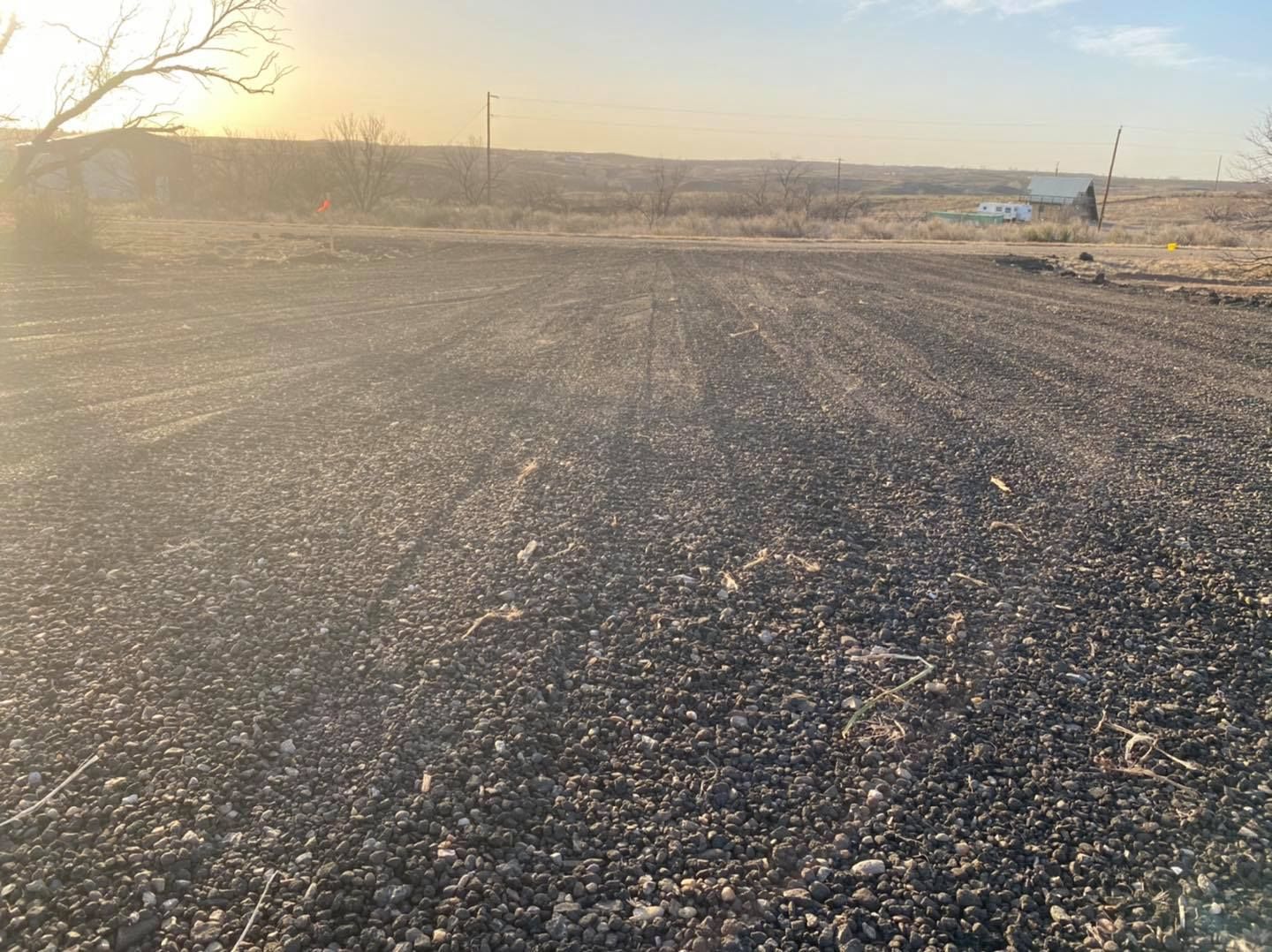 Gravel road in an open field at sunset, with distant buildings and bare trees.
