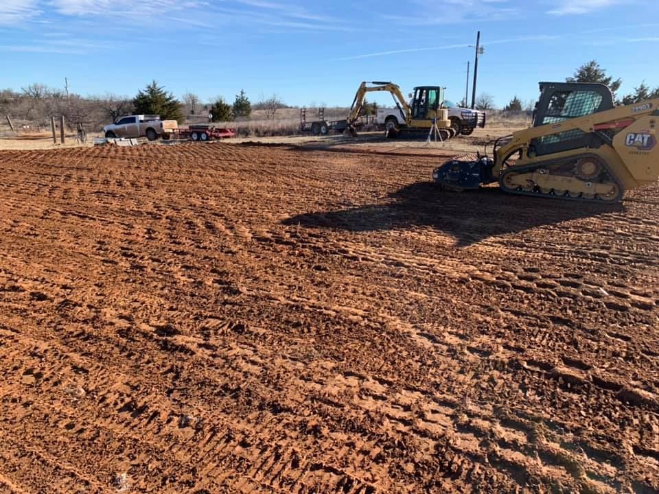 Construction site with red dirt being leveled by a skid steer, with excavators and trucks.