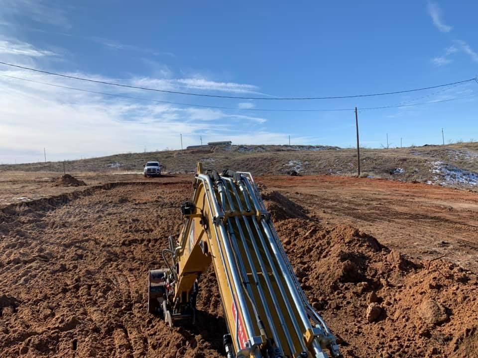 An excavator digging in a dirt field under a blue sky. In the distance, cars sit on a hill.