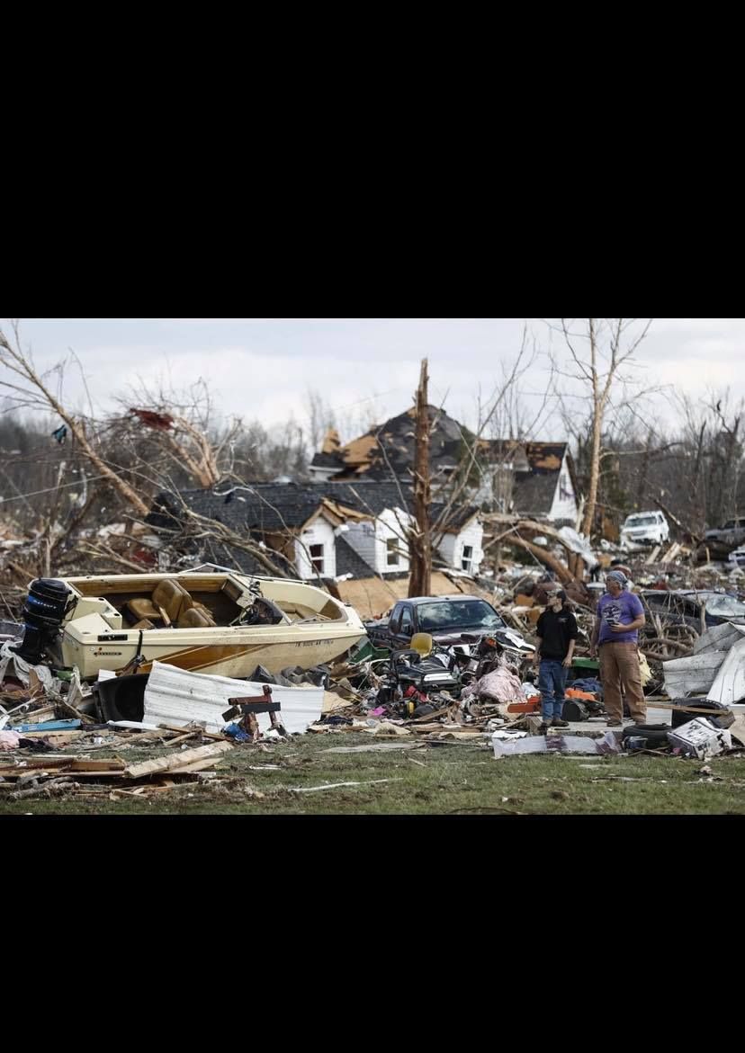 Devastated home and yard, debris scattered. Boat and cars wrecked, two people standing, trees damaged.