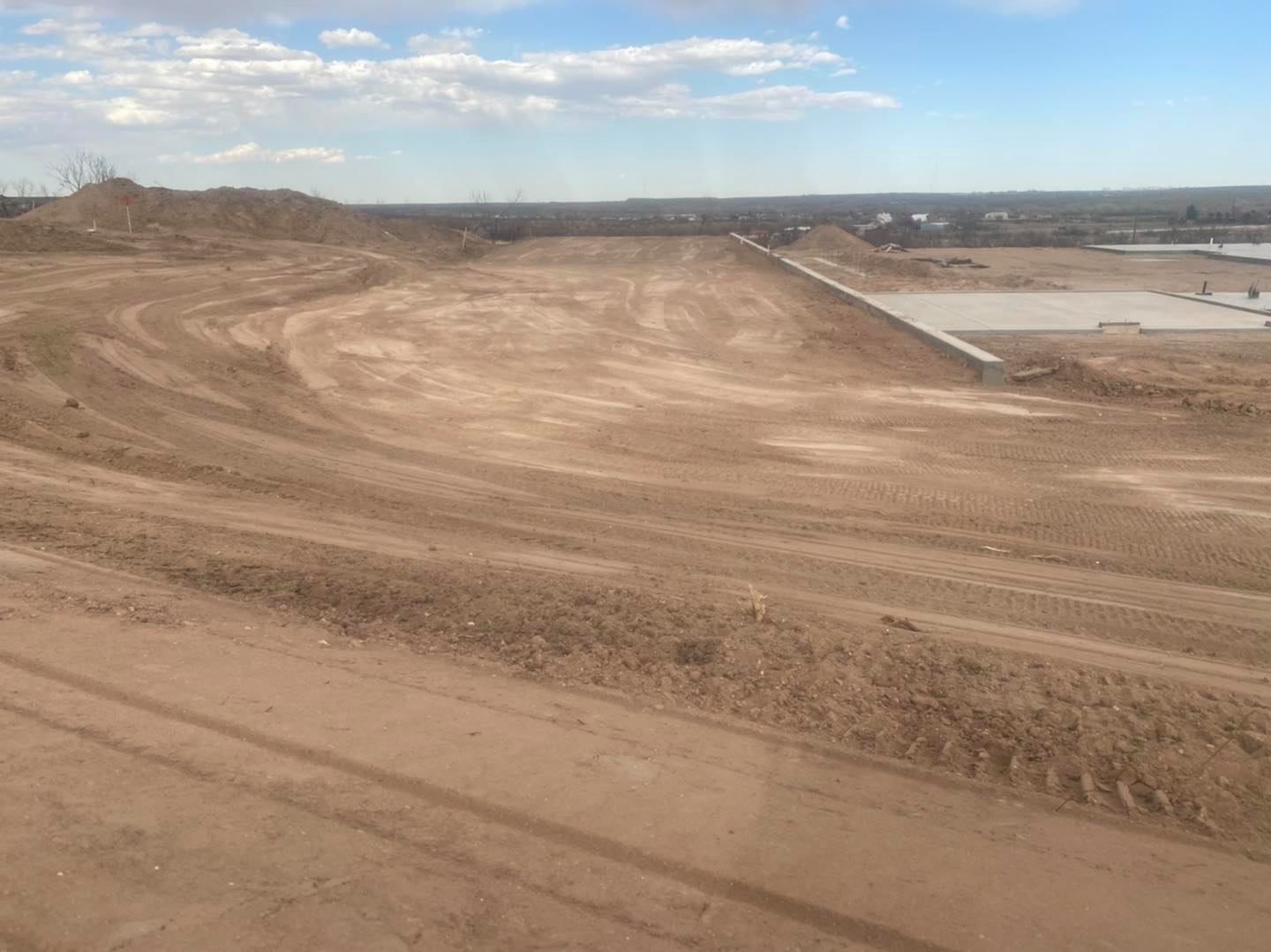 Dirt terrain with tire tracks. Cloudy sky, construction site in the distance.