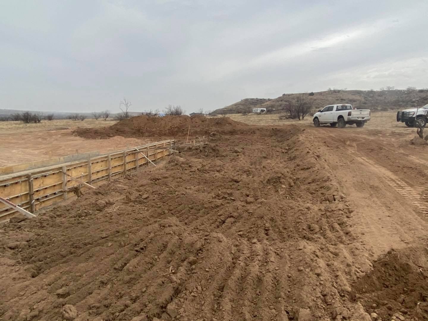 Construction site with dirt mounds, wooden forms, and white pickup trucks.