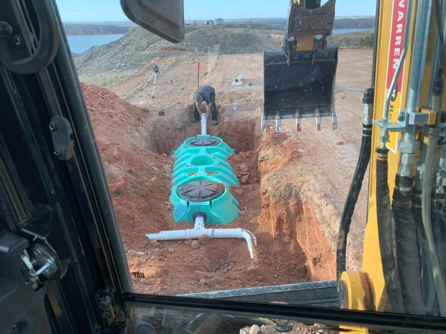 Excavator lowering a teal septic tank into a trench dug in red dirt.