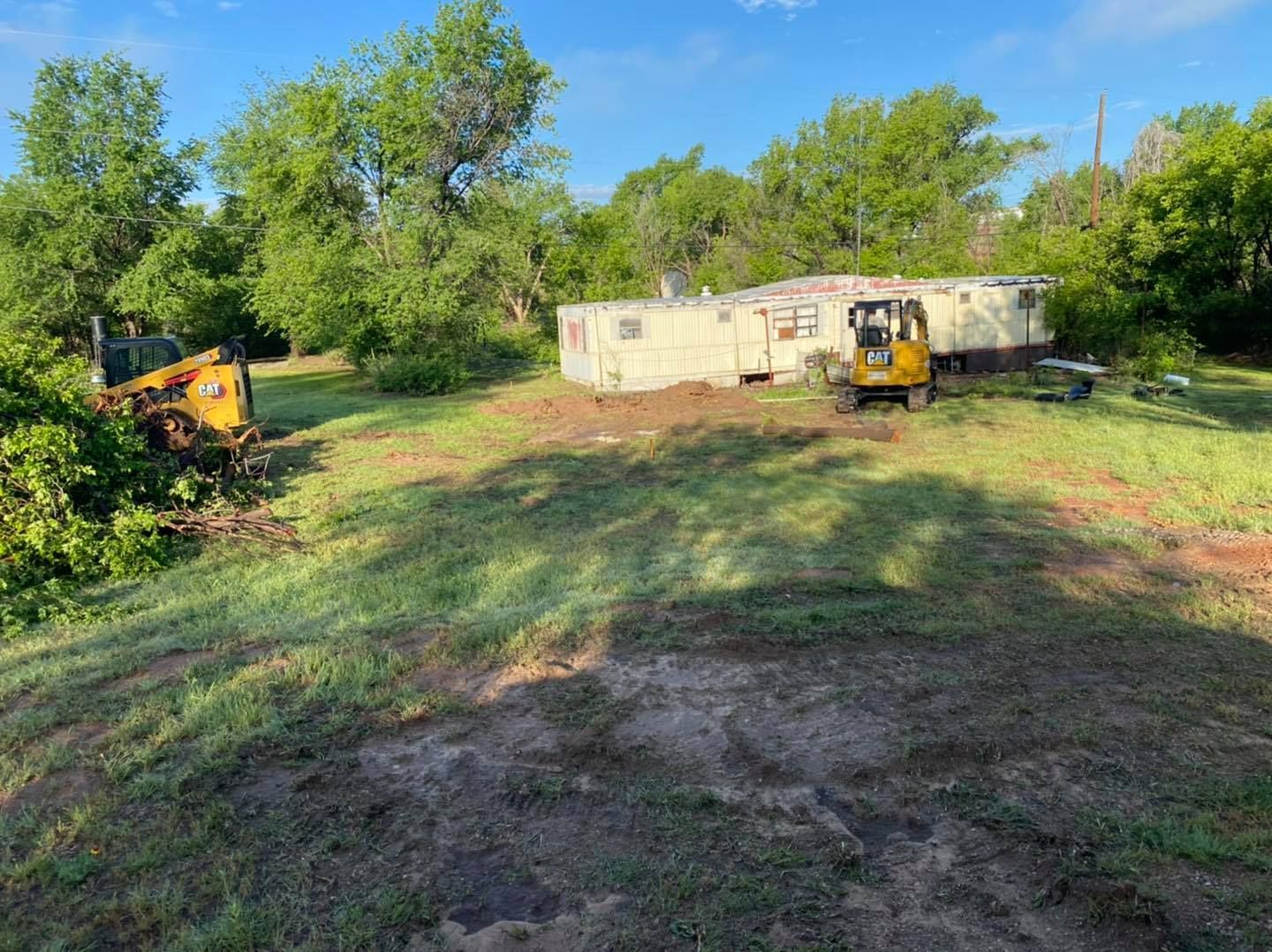 Two construction vehicles and a dilapidated house on a grassy lot. Bright daylight, blue sky.