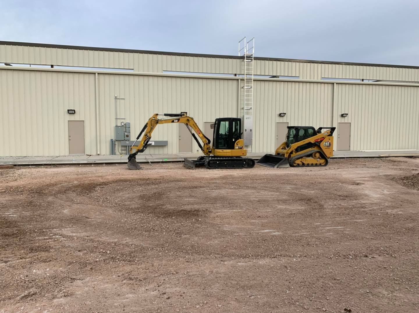 A small excavator and a skid steer in front of a tan building on a gravel lot.