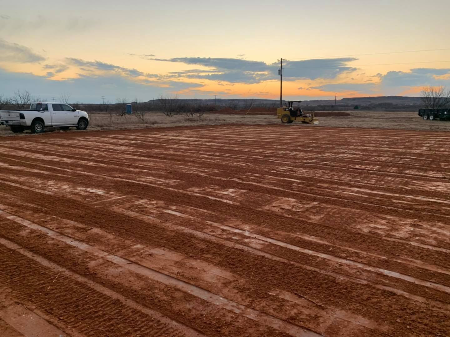 Construction site with a truck, compactor, and sunset over red dirt.