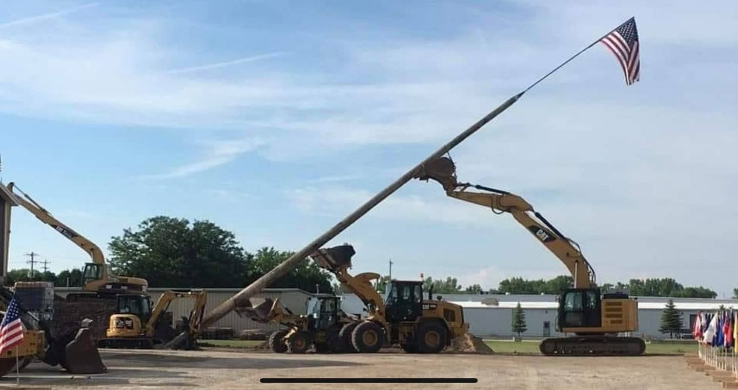 Construction equipment displaying American flag. Excavator holding long pole with flag against blue sky.
