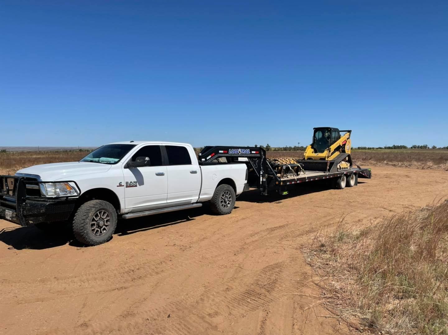 White pickup truck towing a trailer with a yellow skid steer on a dirt road under a blue sky.