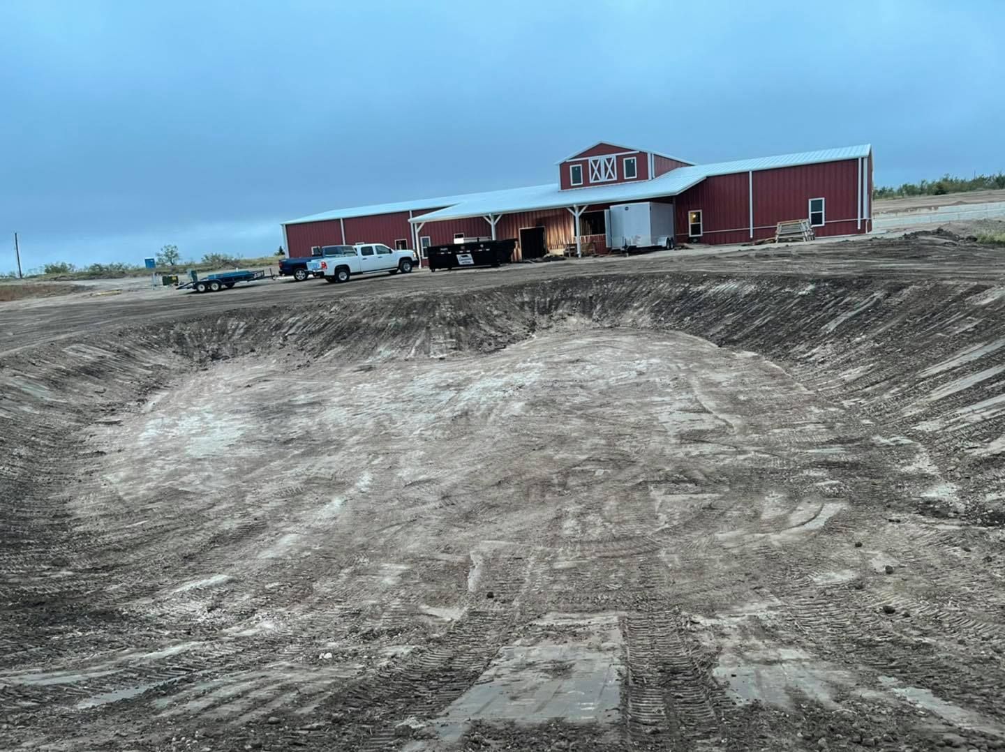 Construction site with red building, dug-out earth, and overcast sky. Trucks parked nearby.