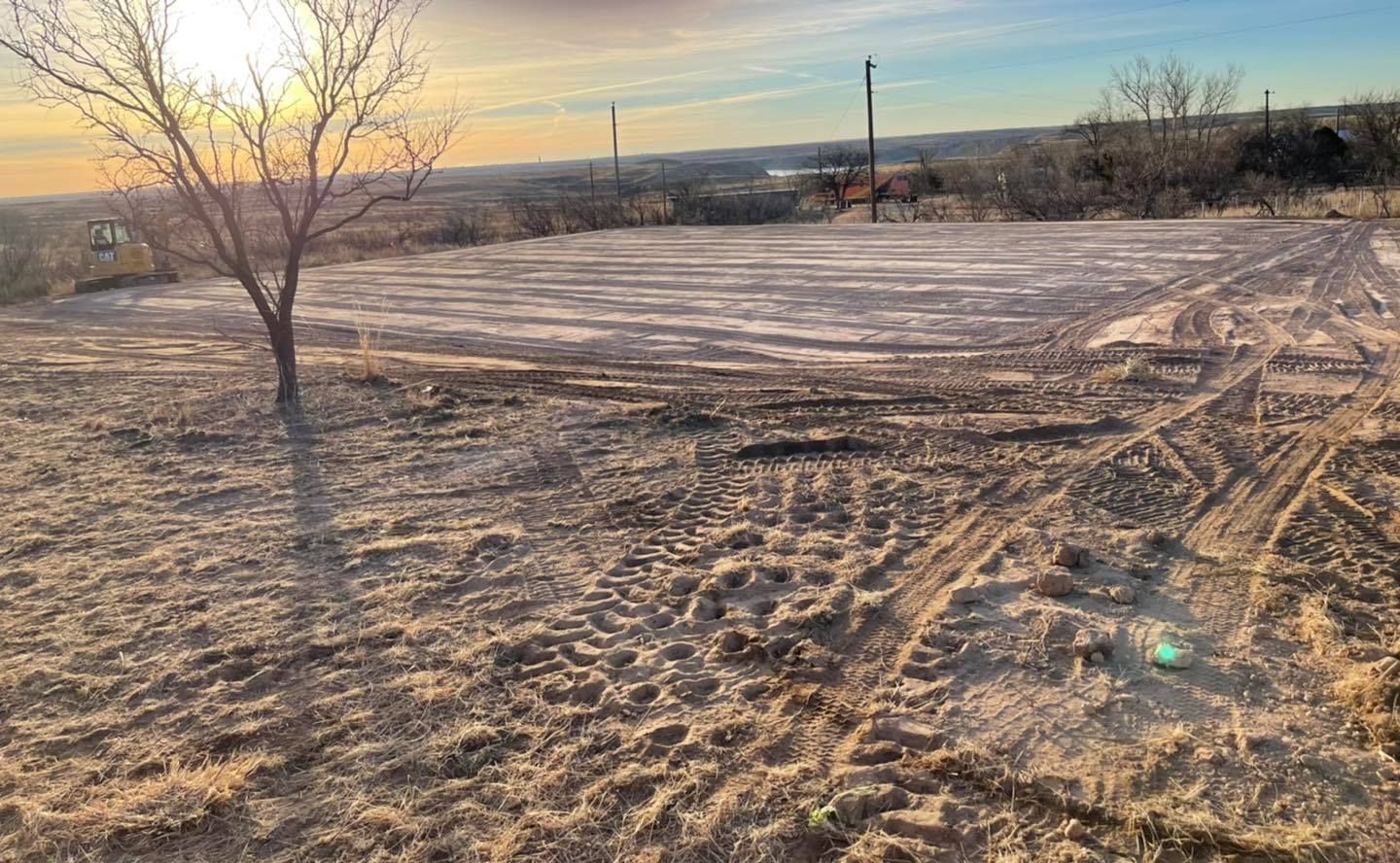 Bare ground with tire tracks, a bare tree, and a distant view under a sunset sky.