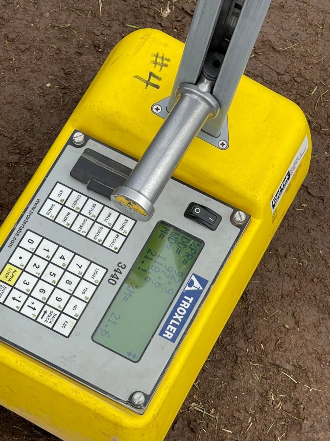 Yellow Troxler nuclear density gauge on soil; shows readings on screen.