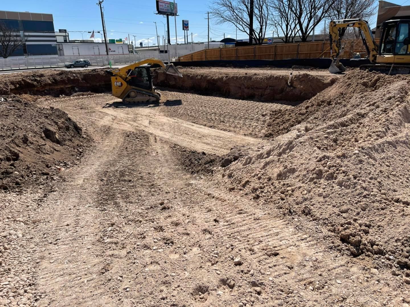 Construction site with an excavator digging in a dirt pit. Brown and tan earth dominates.