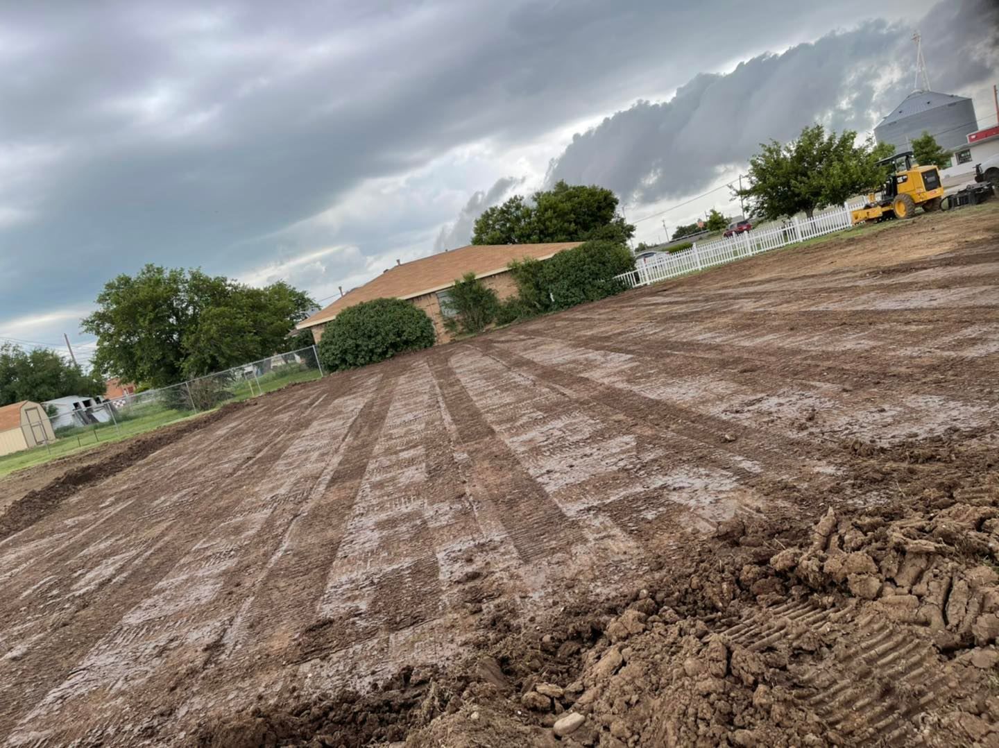 Muddy dirt field with rows, overcast sky, houses and a yellow excavator in the background.
