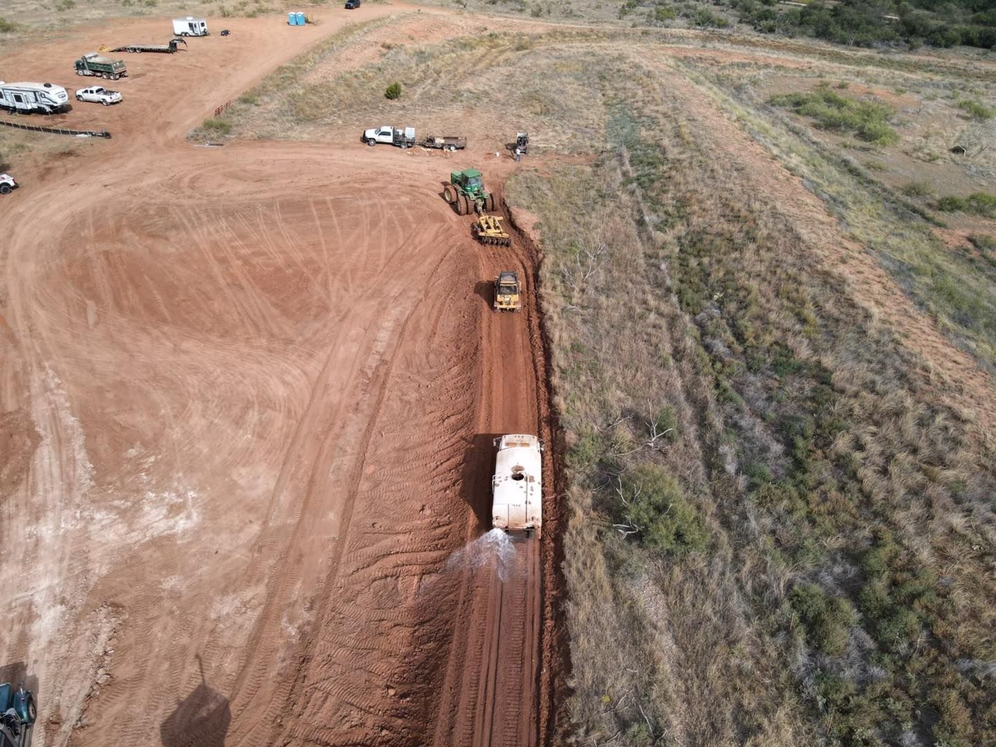 Aerial view of construction site; truck spraying water on red dirt road, bulldozers, and trailers nearby.