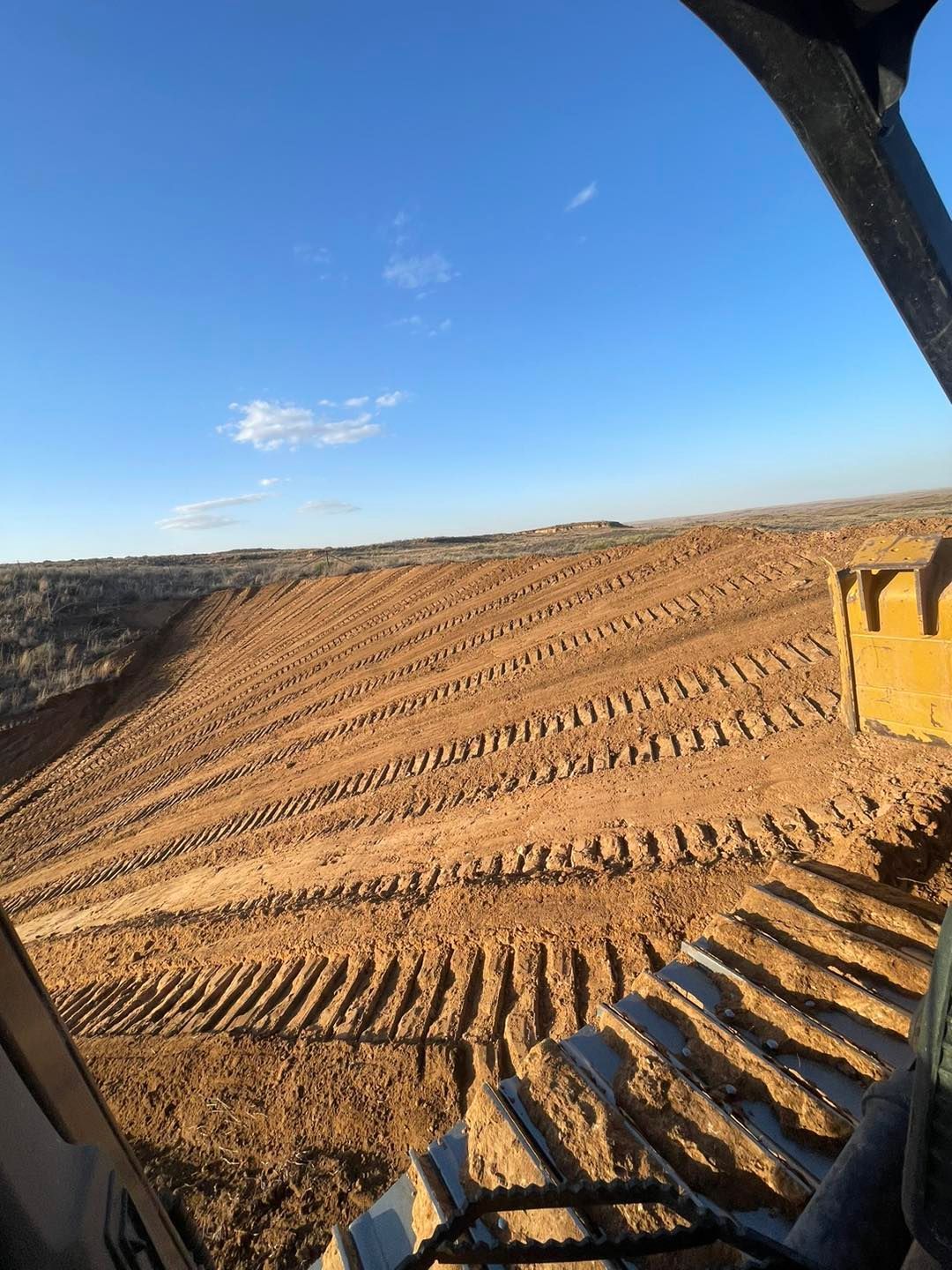 View from an excavator cab shows freshly graded brown earth under a blue sky.