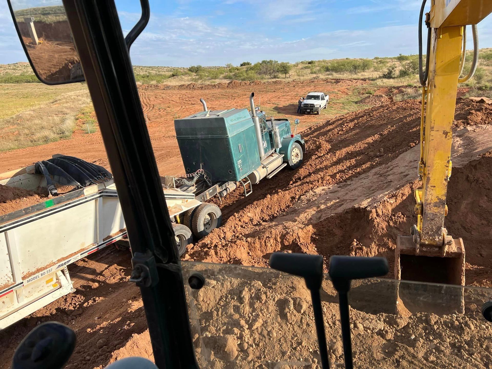 A view from an excavator cab showing a green semi-truck and a white pickup parked on a red dirt construction site.