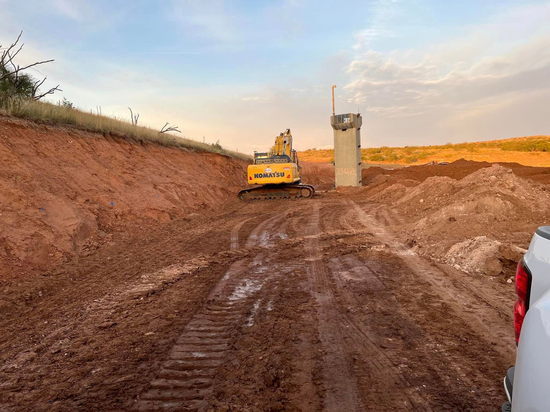 Excavator in muddy terrain beside a concrete pillar, earthworks in progress outdoors.