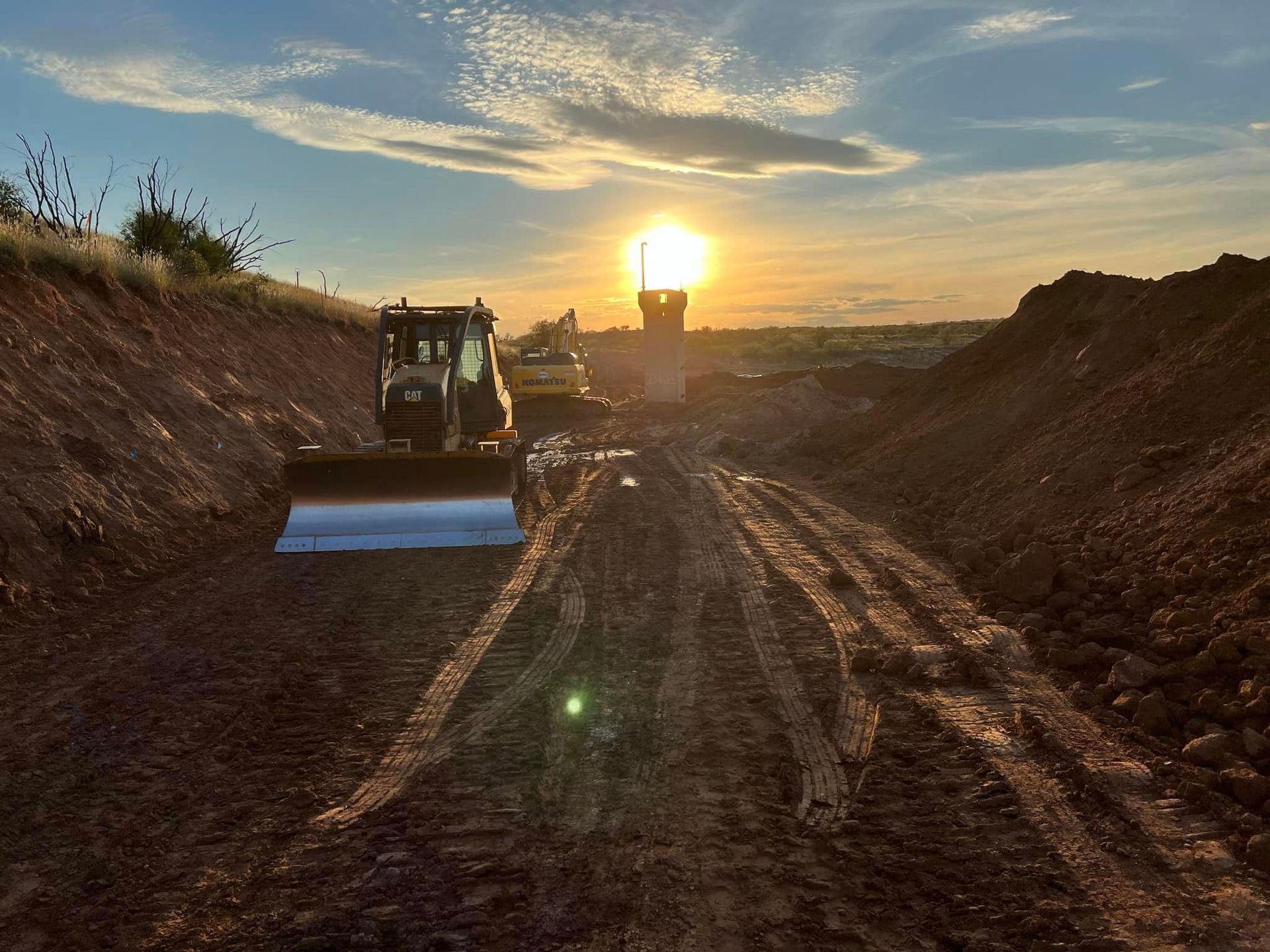 Construction equipment at sunset in a trench; dirt paths lead towards the setting sun.