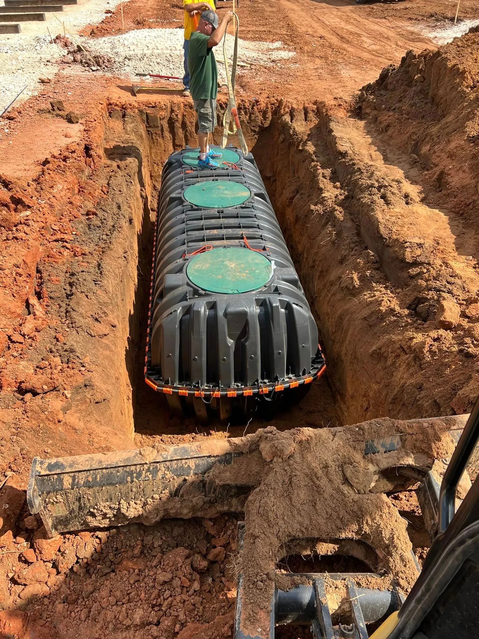 Worker installing black septic tank in a dirt excavation site with a crane.