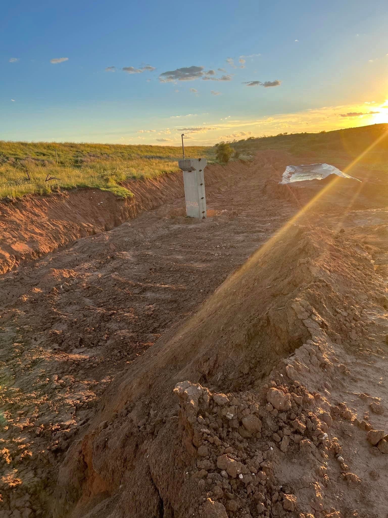 Dirt trench with a concrete post in an open field at sunset.