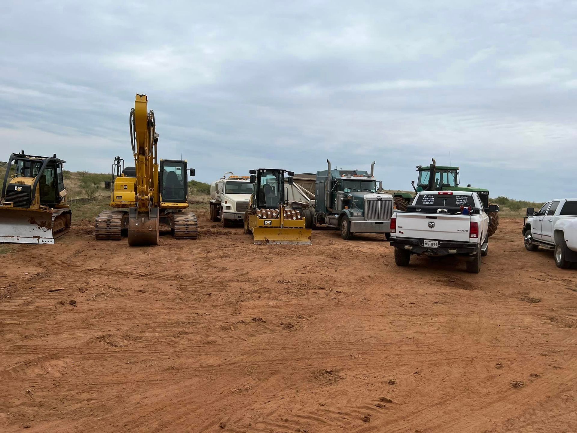 Construction vehicles on a red dirt lot under a cloudy sky.