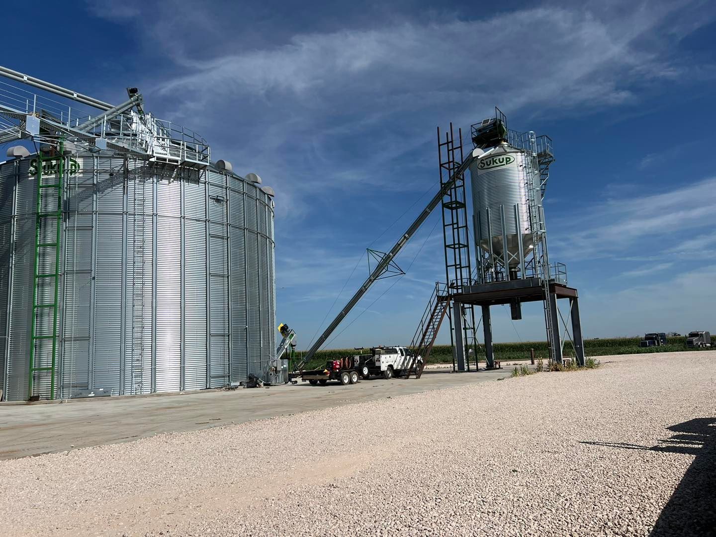 Large silver grain silo and smaller elevated silo, connected by a conveyor belt, outdoors under a blue sky.