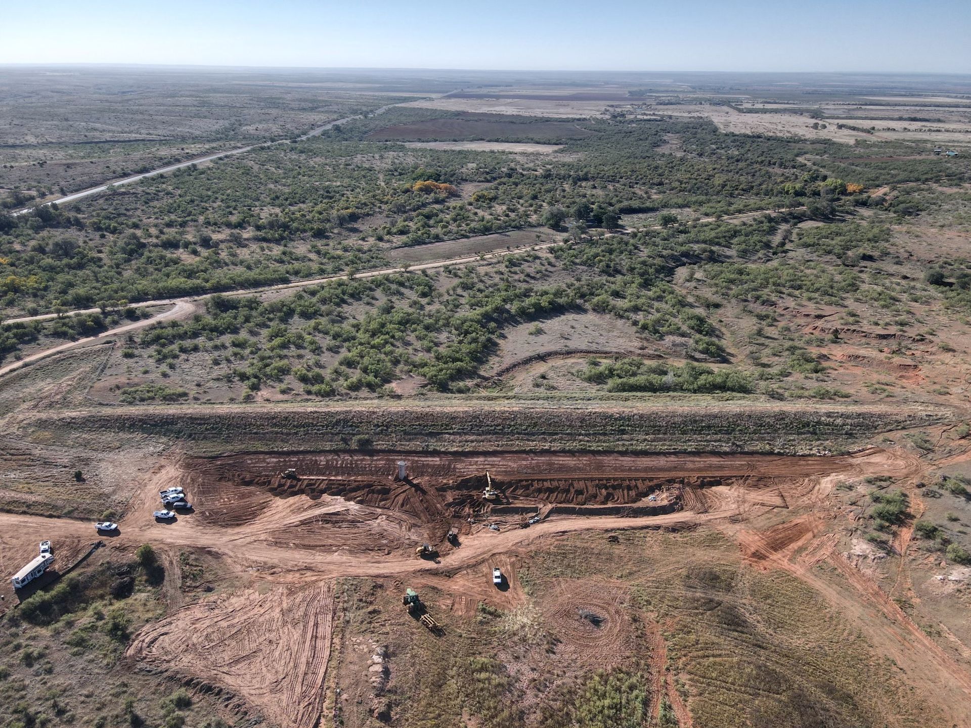 Aerial view of construction site in a dry, grassy landscape; earth-moving equipment works near a long, earthen dam.