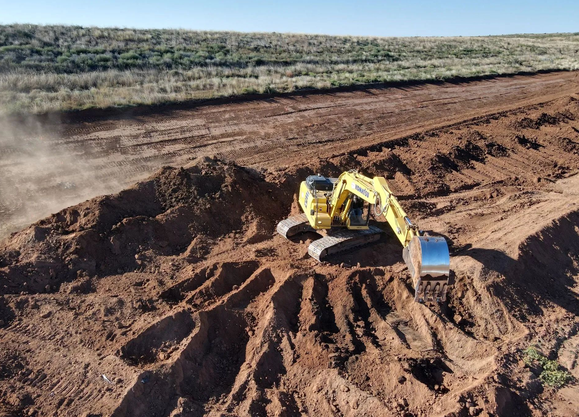 Yellow excavator digging red dirt in a wide construction site under a clear sky.
