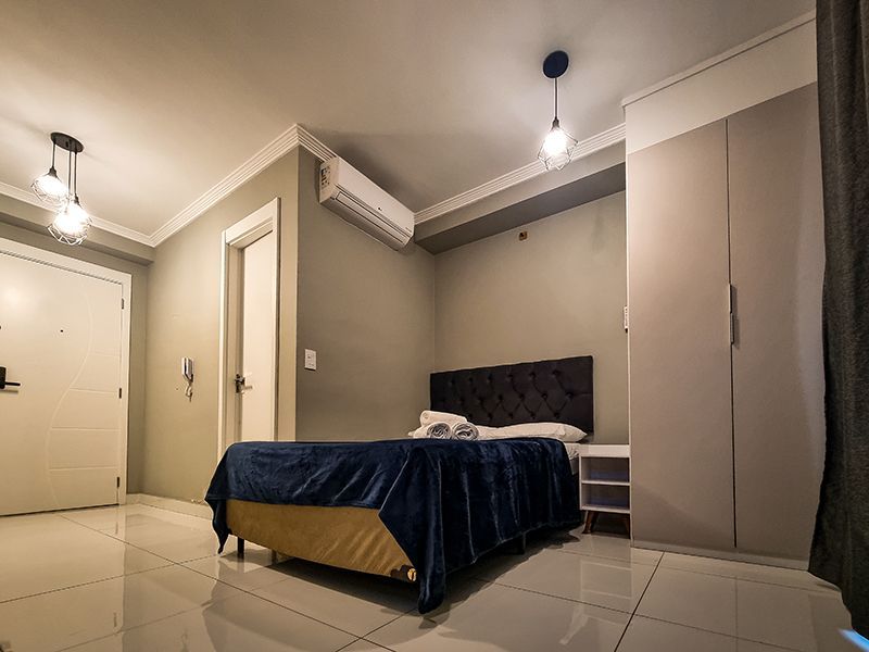 Quarto moderno com cama, piso de azulejo, paredes bege e luminárias pendentes no teto.